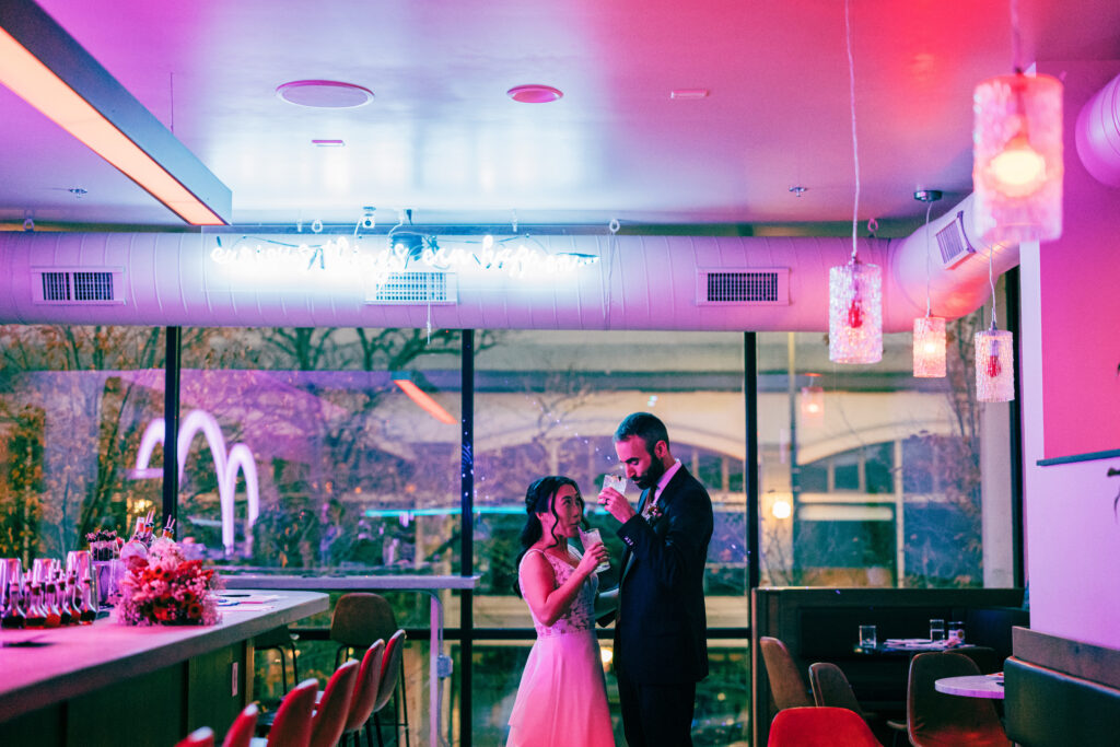 Bride and groom sipping cocktails together in a neon-lit bar with pink and blue ambient lighting.