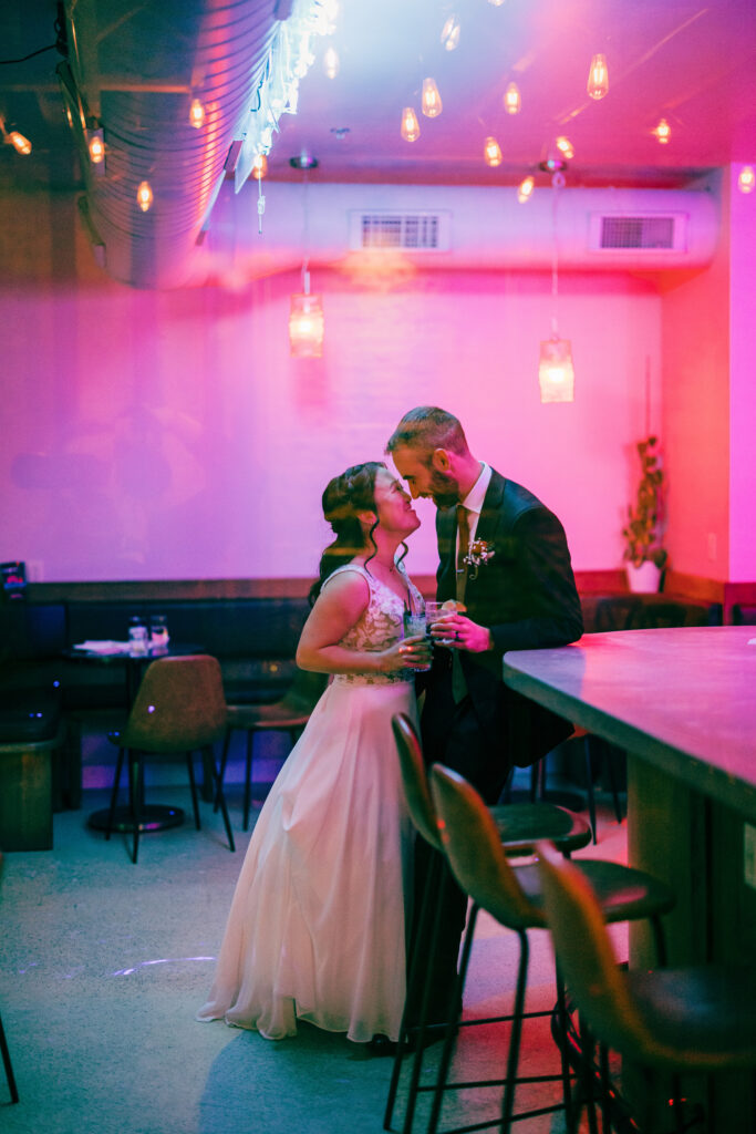 Bride and groom holding cocktails and embracing beside a bar under warm pink neon lighting.