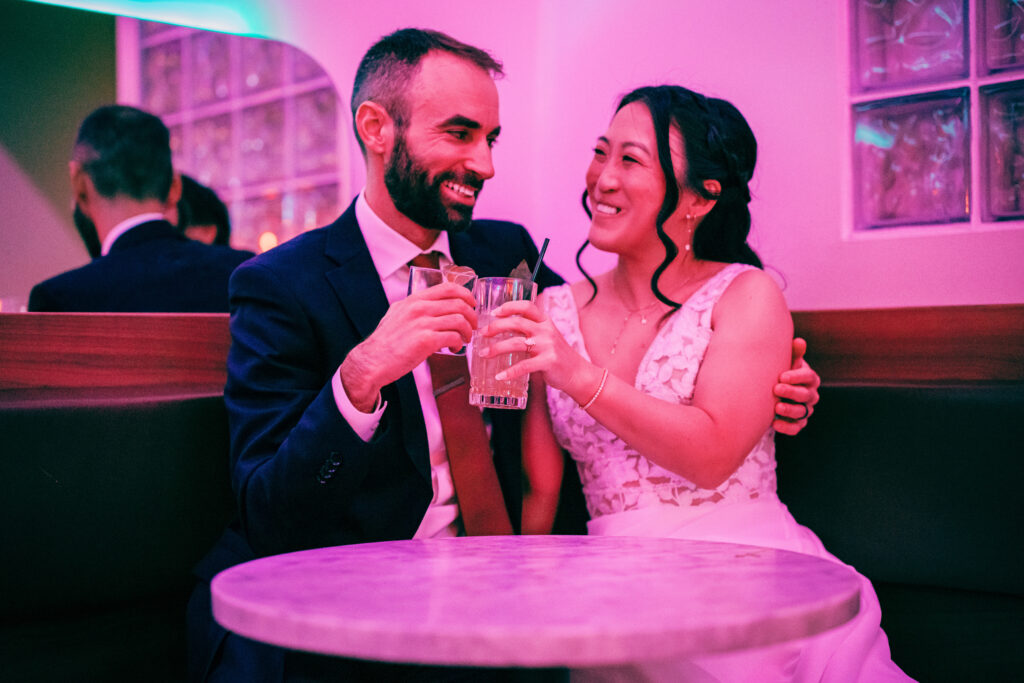Bride and groom toasting cocktails together in a cozy neon-lit cocktail lounge after their wedding.