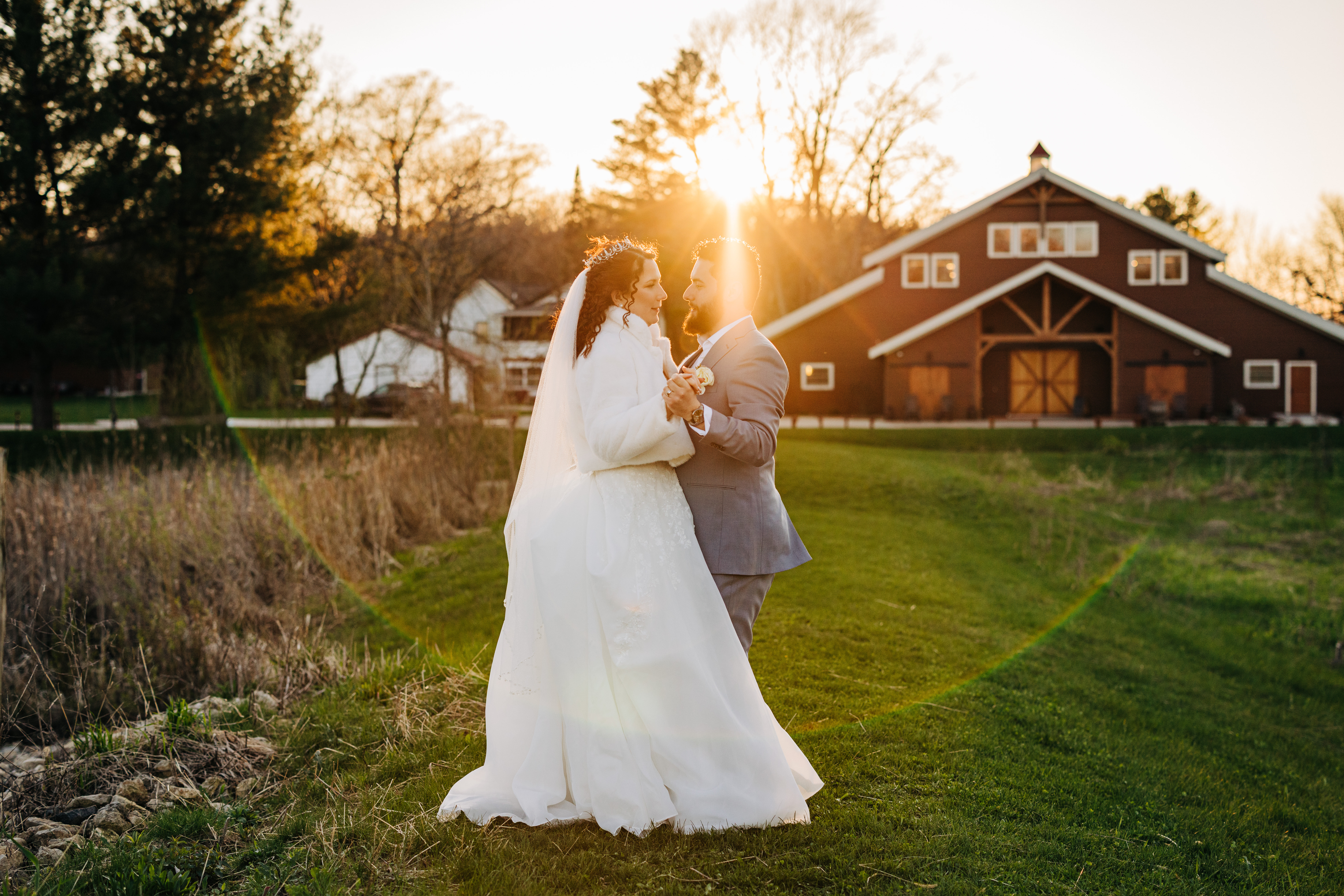 Bride and groom share a sunset dance outside a rustic barn wedding venue with golden backlight.