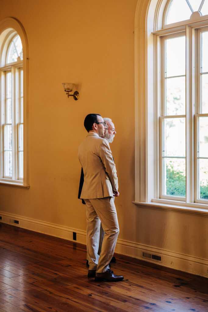 Groom and guest standing beside tall arched windows before the wedding ceremony.