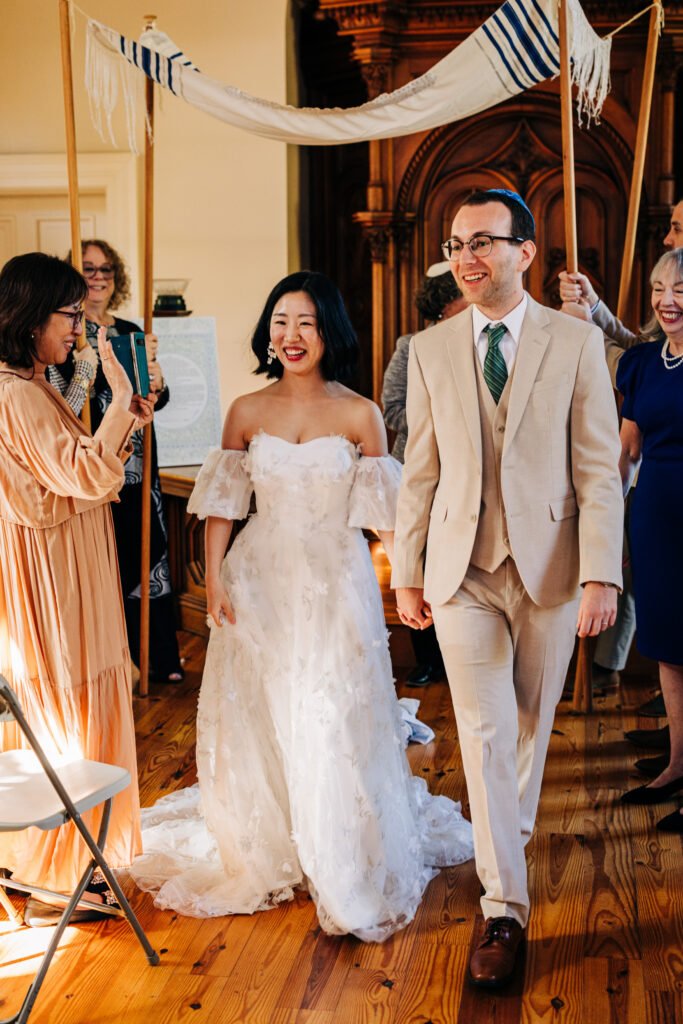 Bride and groom walk beneath a chuppah during an intimate indoor wedding ceremony surrounded by guests.