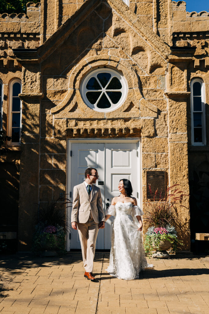 Bride and groom hold hands outside a historic stone building with arched windows and sunlit architecture.