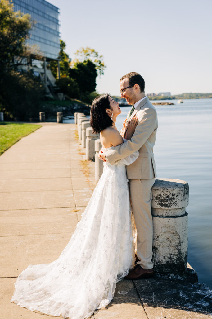 Bride and groom embrace along a waterfront promenade with calm blue water in the background.