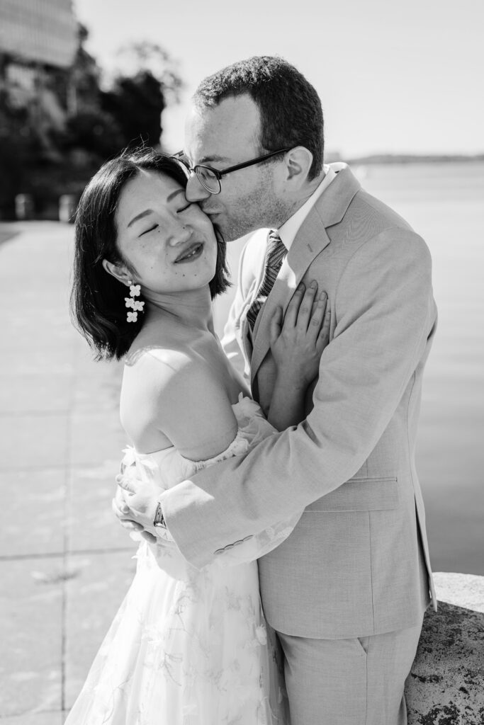 Black-and-white portrait of a groom kissing the bride’s cheek beside a waterfront walkway.