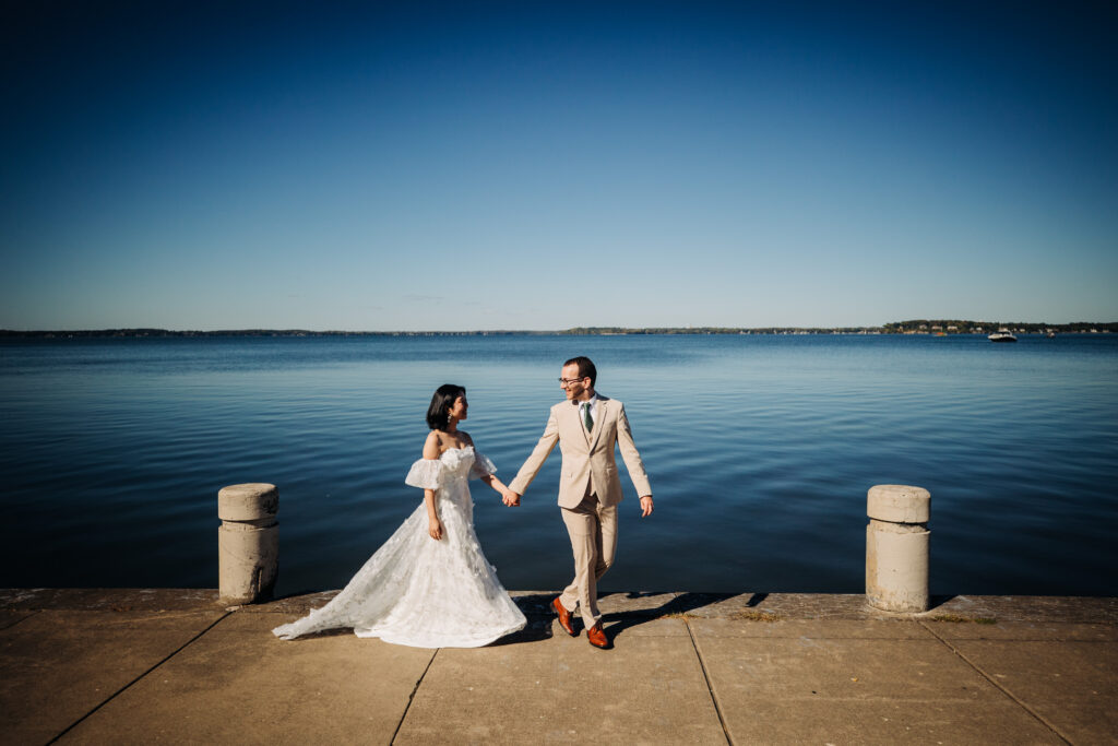 Bride and groom walk hand in hand along a waterfront pier beneath a clear blue sky.