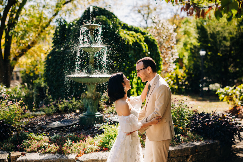 Bride and groom stand together near a sparkling garden fountain surrounded by colorful flowers.