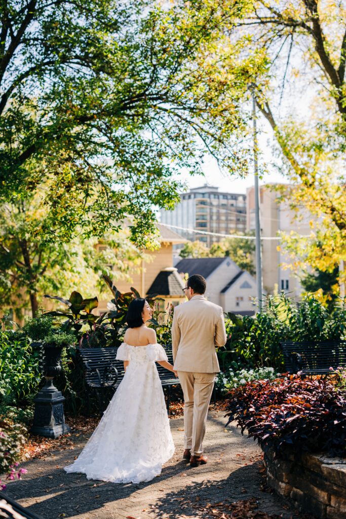 Bride and groom walk hand in hand along a shaded garden path framed by trees and flowers.