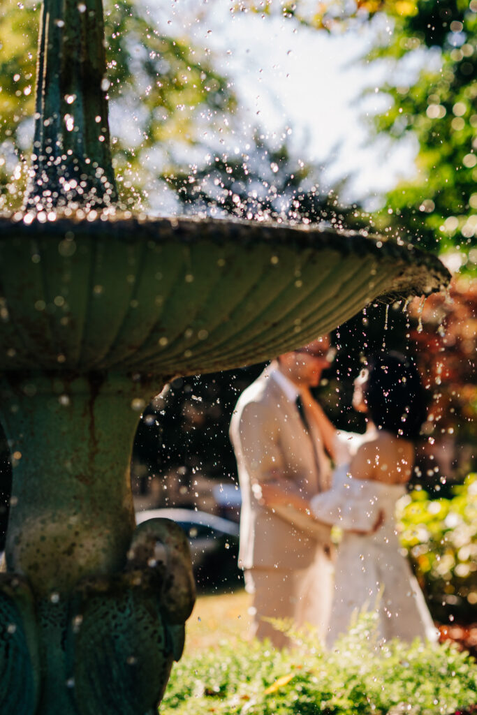 Water droplets sparkle in the foreground as a bride and groom embrace in a garden beyond.