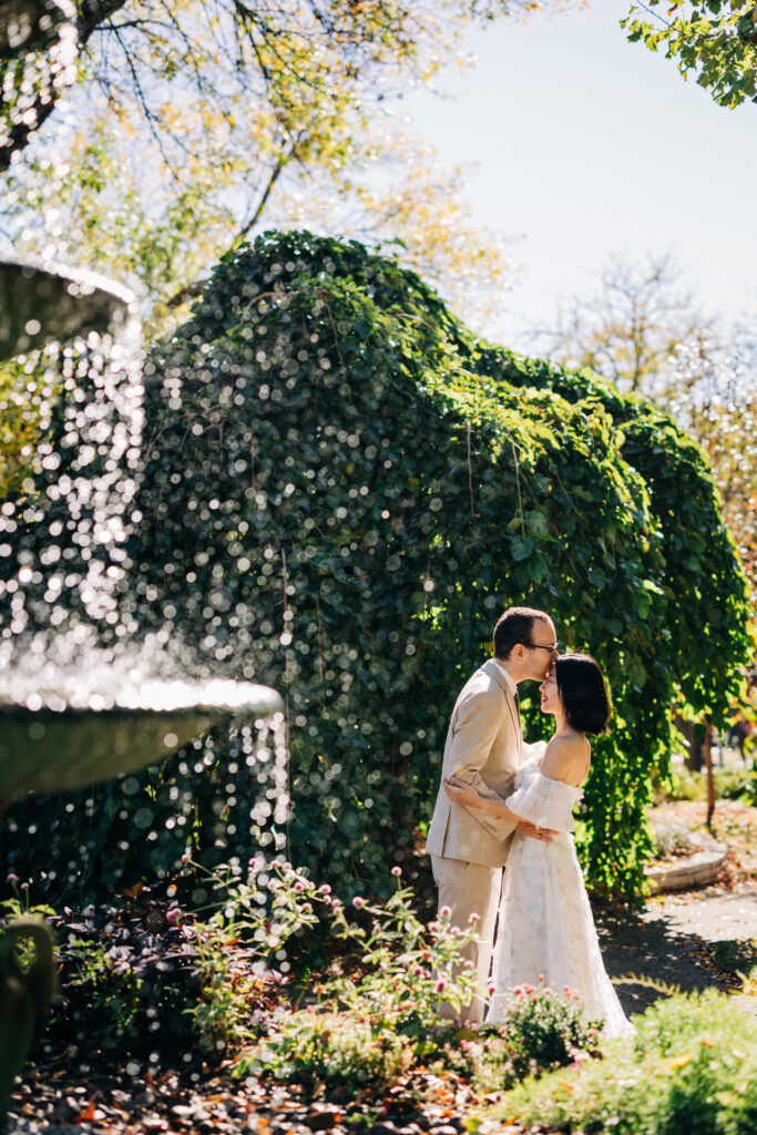 Bride and groom share a kiss beside a garden fountain framed by cascading greenery and sunlight.