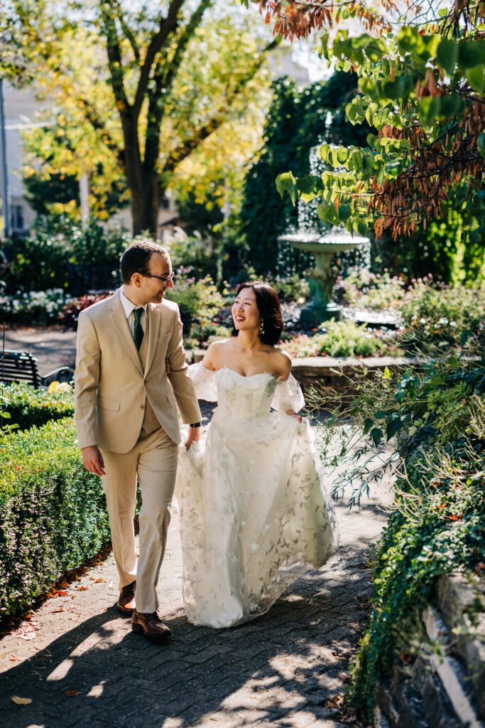 Newlyweds walk through a lush garden pathway with a fountain and flowering plants nearby.