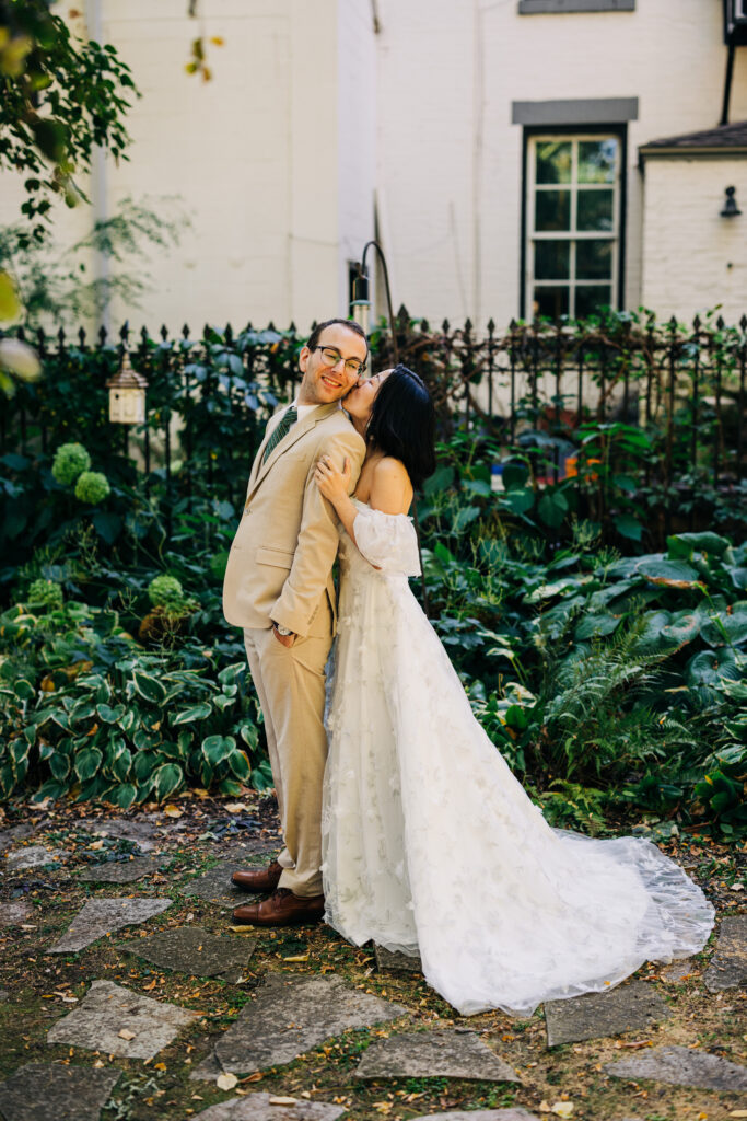 Bride in a floral wedding gown embraces groom in a beige suit during an outdoor garden wedding portrait.
