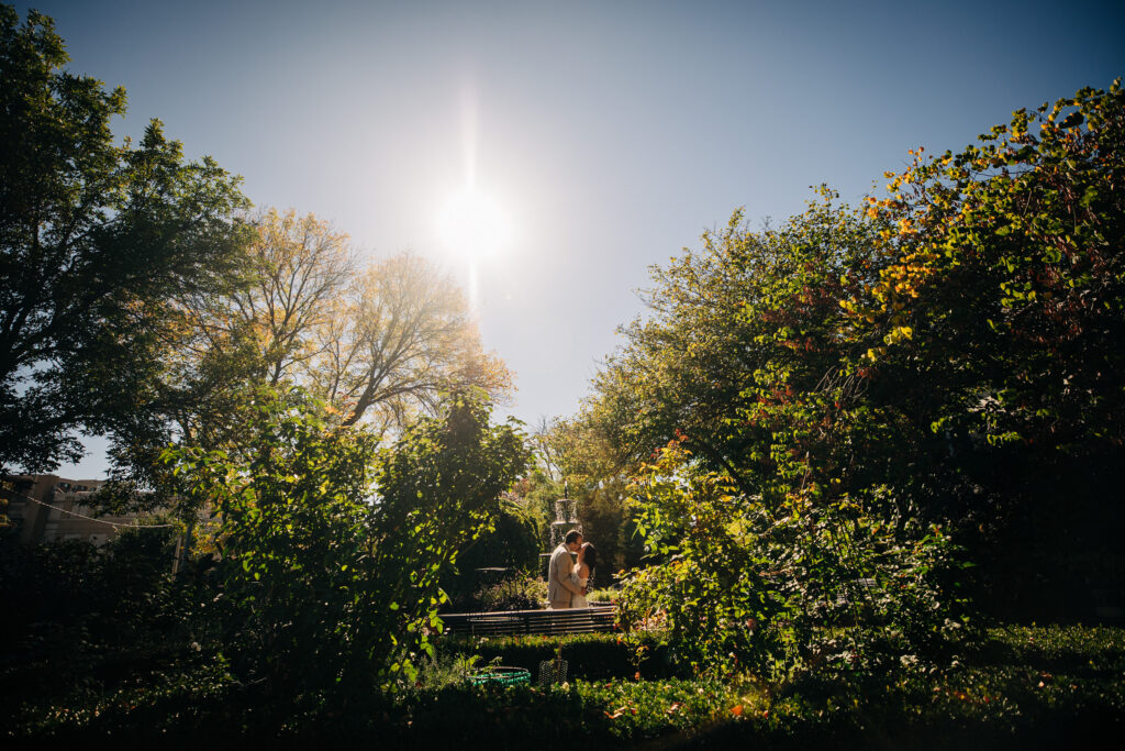 Bride and groom share a quiet moment in a sunlit garden surrounded by lush greenery and autumn foliage.