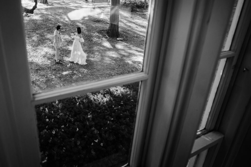 Bride and groom standing together in a garden, viewed from above through a large window.