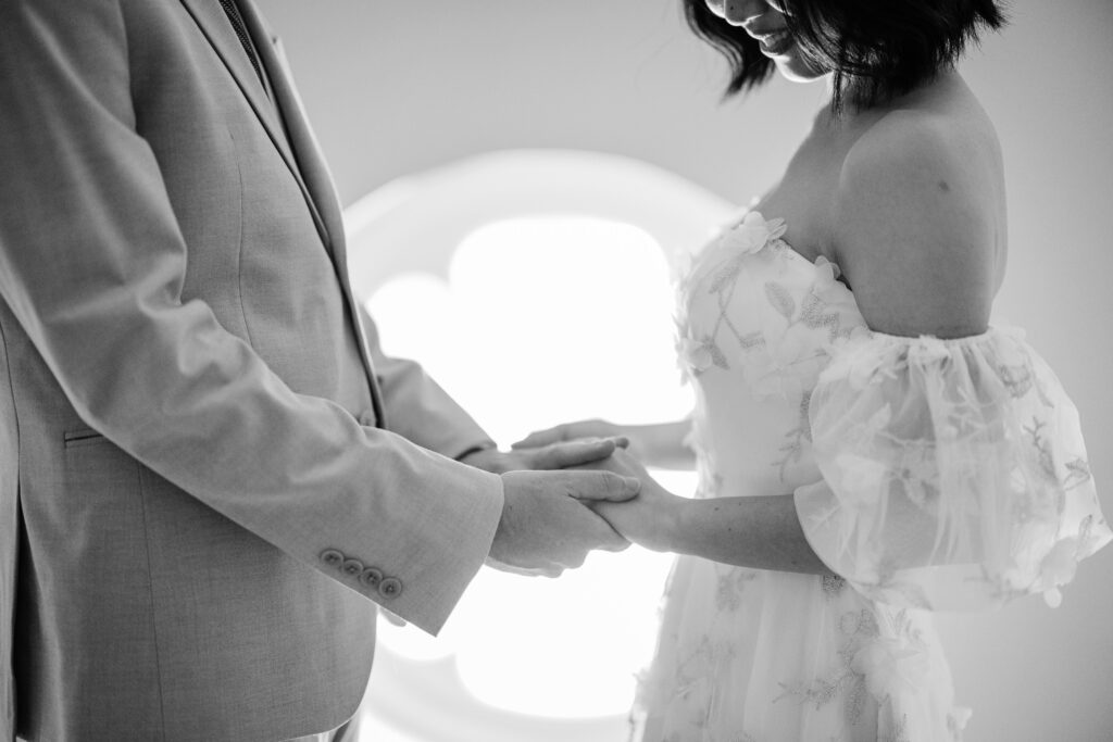 Black-and-white close-up of bride and groom holding hands in front of a softly lit window.