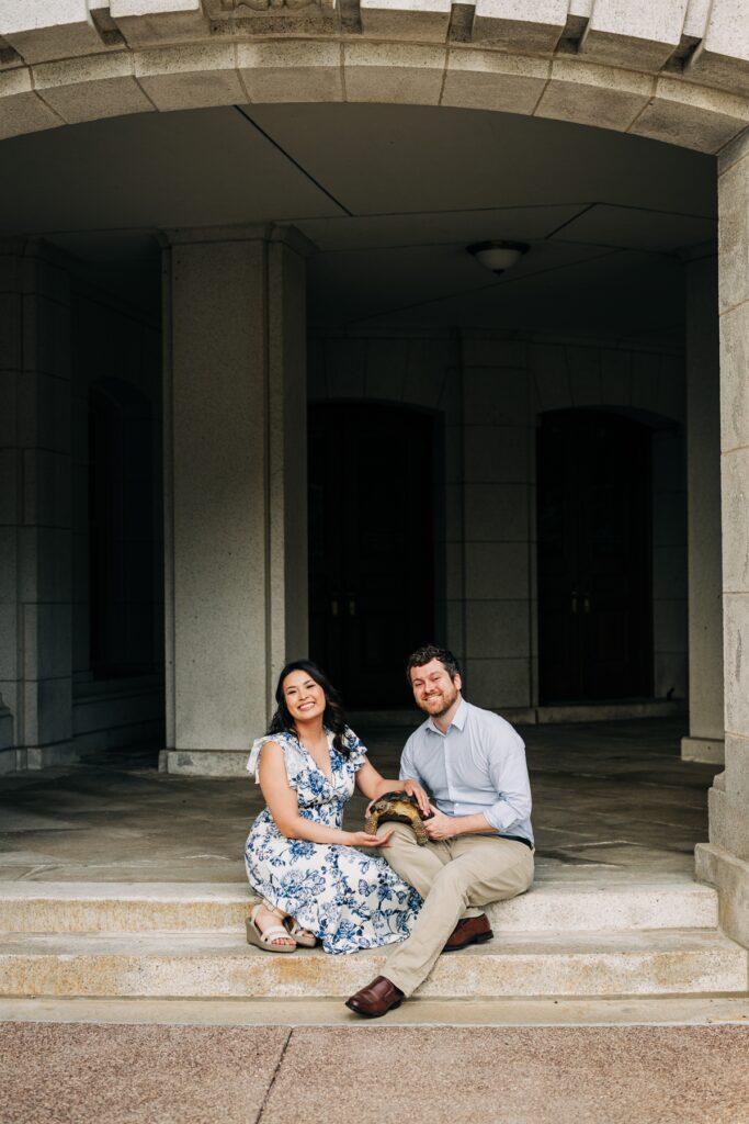 Couple sitting on stone steps with their tortoise beneath a historic archway in downtown Madison during an engagement session.
