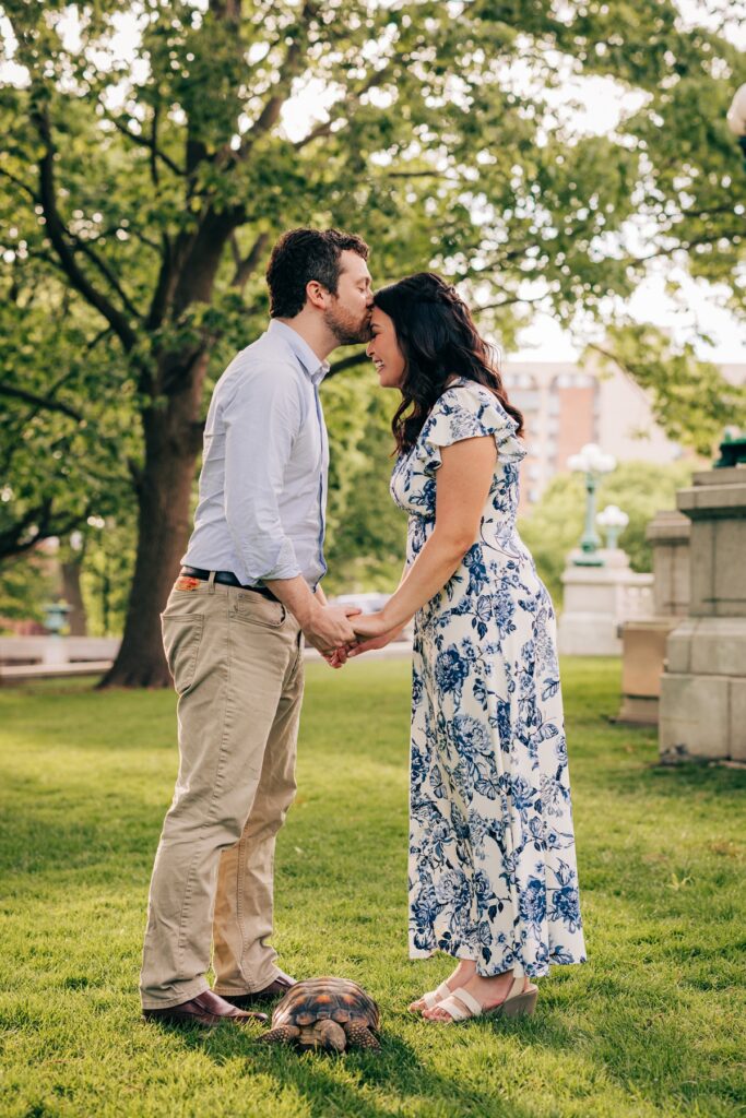 Groom-to-be kisses bride-to-be's forehead as they hold hands with their pet tortoise in the foreground.
