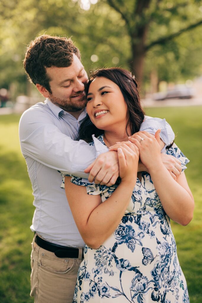 Groom-to-be hugs his fiancée from behind as they smile together in a sunlit park engagement session.
