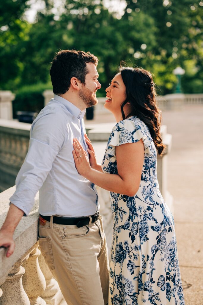 Couple smiling at one another while leaning against a stone railing in warm evening light.
