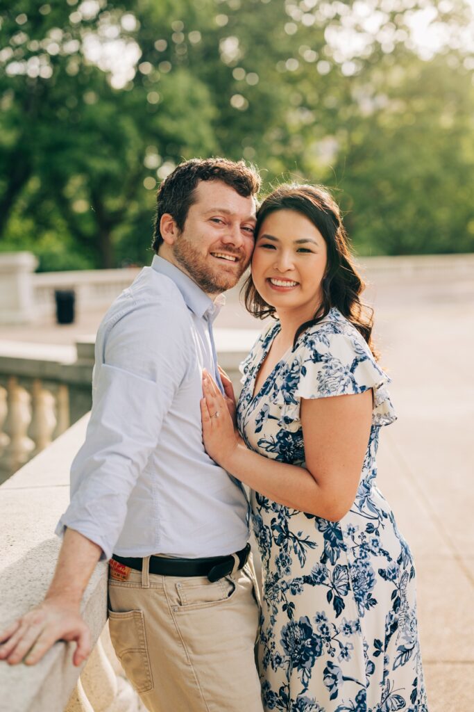 Engaged couple smiling at the camera while embracing beside a stone balustrade at sunset.
