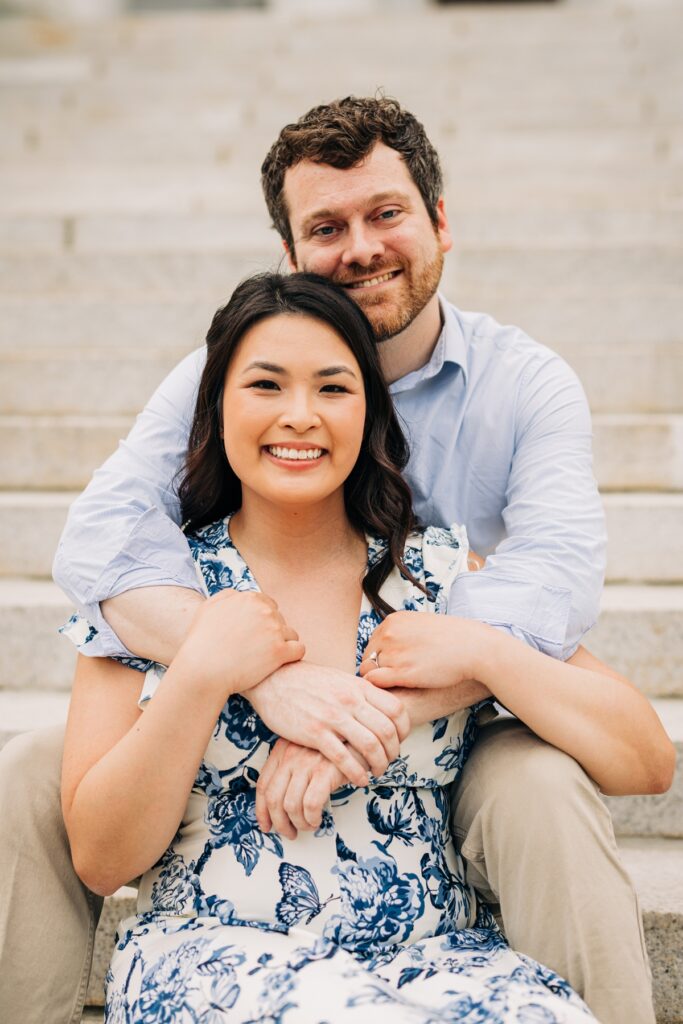 Engaged couple sitting on stone steps with the groom embracing his fiancée from behind.
