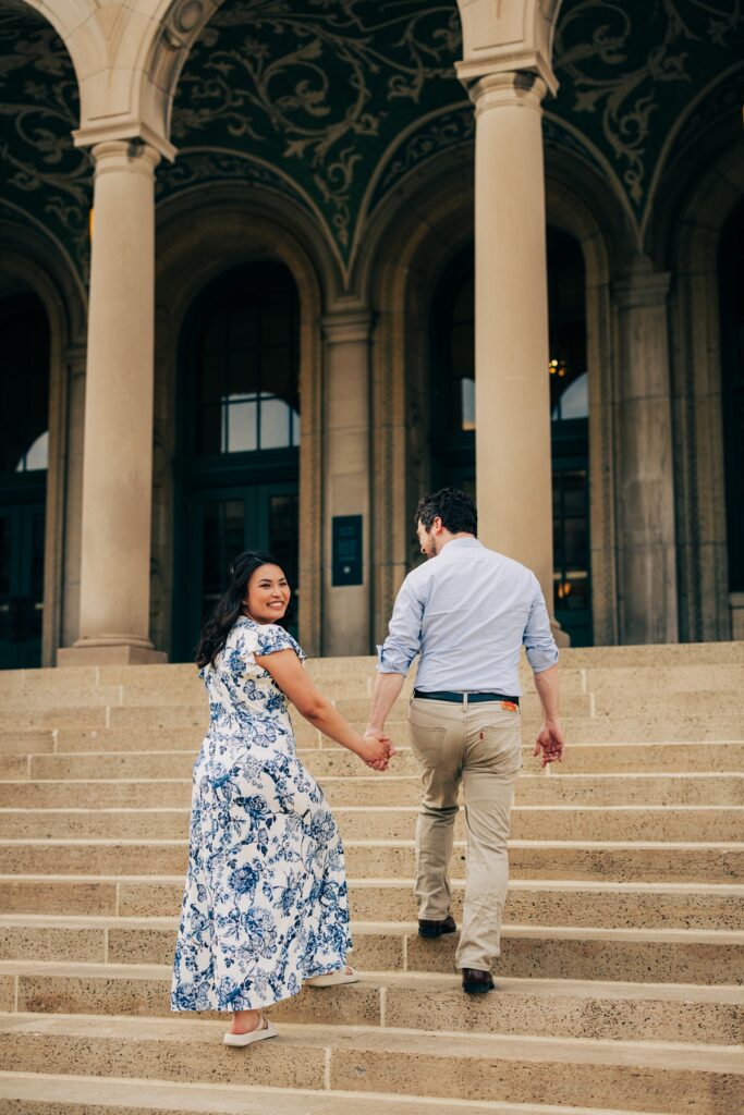Engaged couple walking hand in hand up grand stone steps outside a historic building, looking back with smiles.