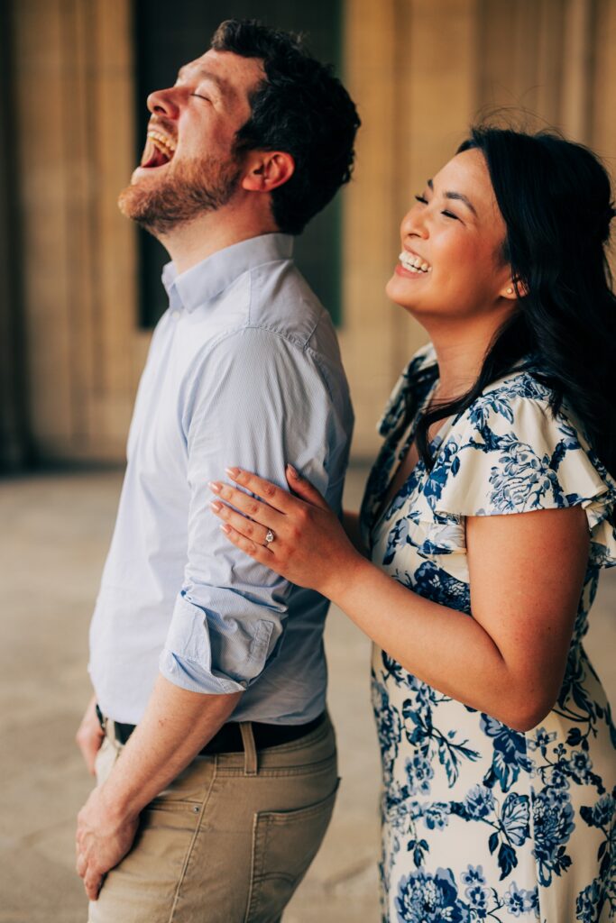 Couple laughing together during an engagement session outside a historic building, with one partner resting a hand on the other's arm.
