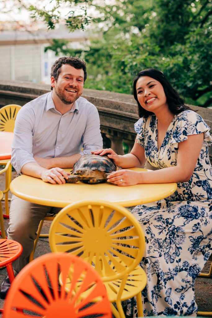 Couple seated at a colorful outdoor café table, smiling while gently petting a tortoise during their engagement session.
