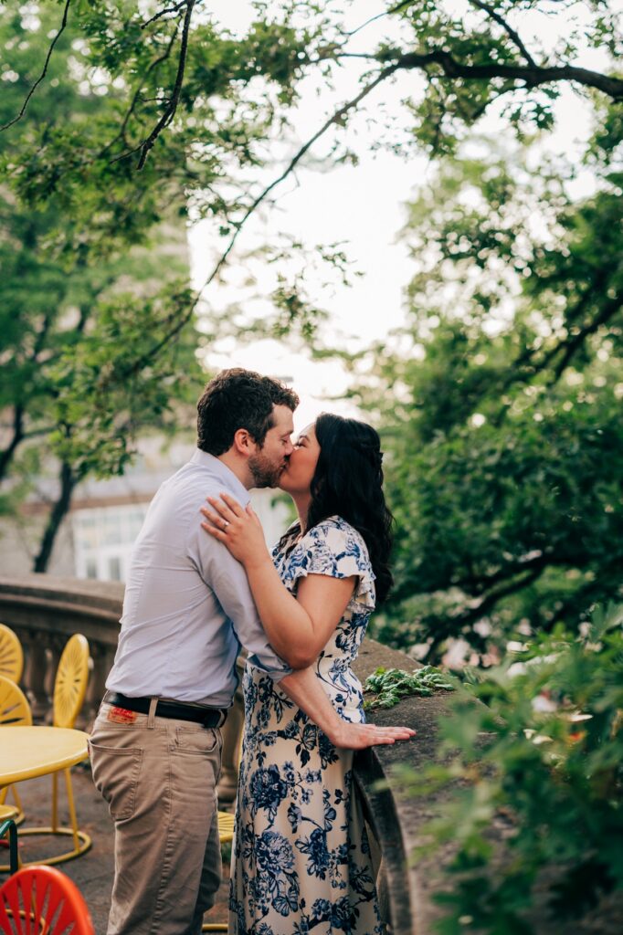 Couple sharing a kiss beside a stone terrace surrounded by lush green trees during an outdoor engagement session.