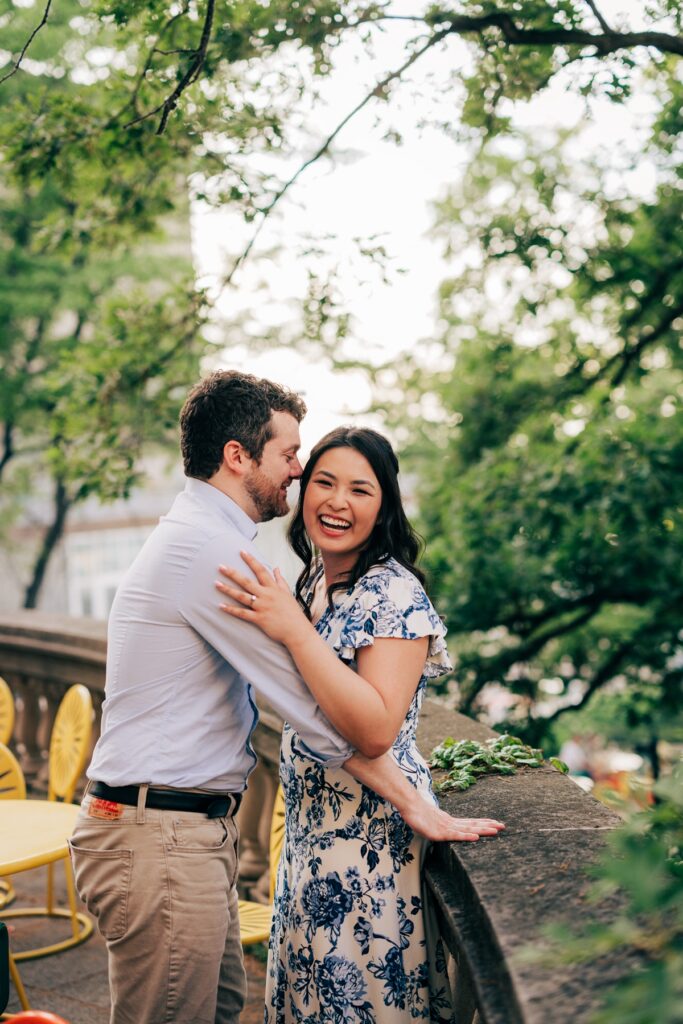 Couple embracing and laughing beside a stone terrace surrounded by greenery during a candid engagement portrait.
