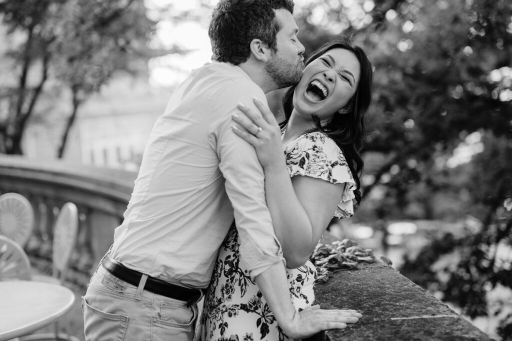 Black-and-white engagement portrait of one partner kissing the other's cheek as they laugh beside a stone terrace.
