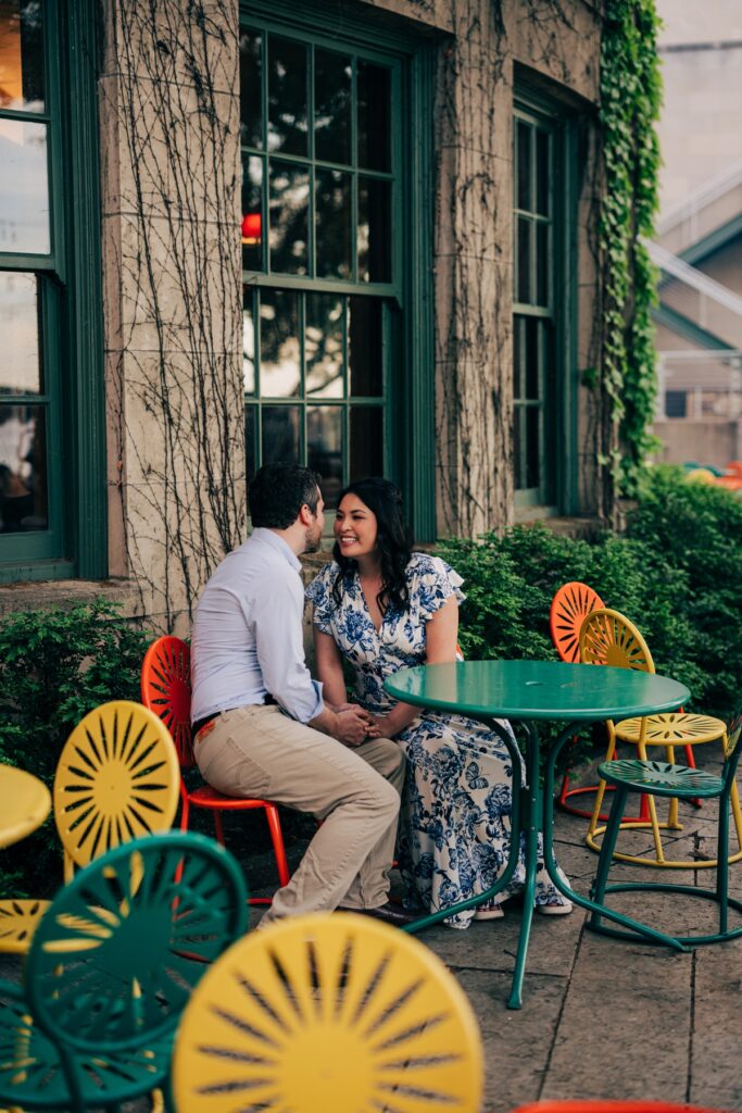Couple sitting together at colorful outdoor café tables, smiling at each other during a relaxed engagement session.

