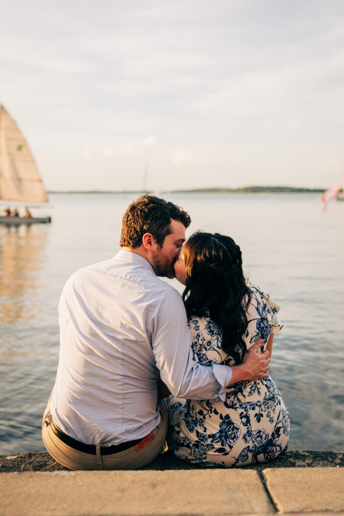 Couple sharing a kiss while sitting along a waterfront at sunset, with sailboats in the background.
