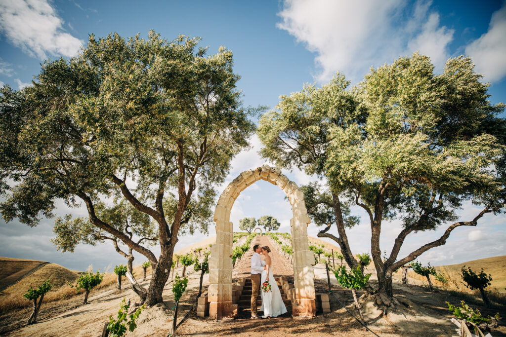 Bride and groom share a kiss beneath a stone arch framed by oak trees at Shandon Chapel Hill overlooking the vineyard.
