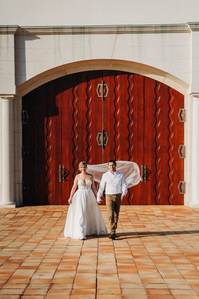 Bride and groom walk hand in hand across the chapel courtyard with the large wooden doors behind them.