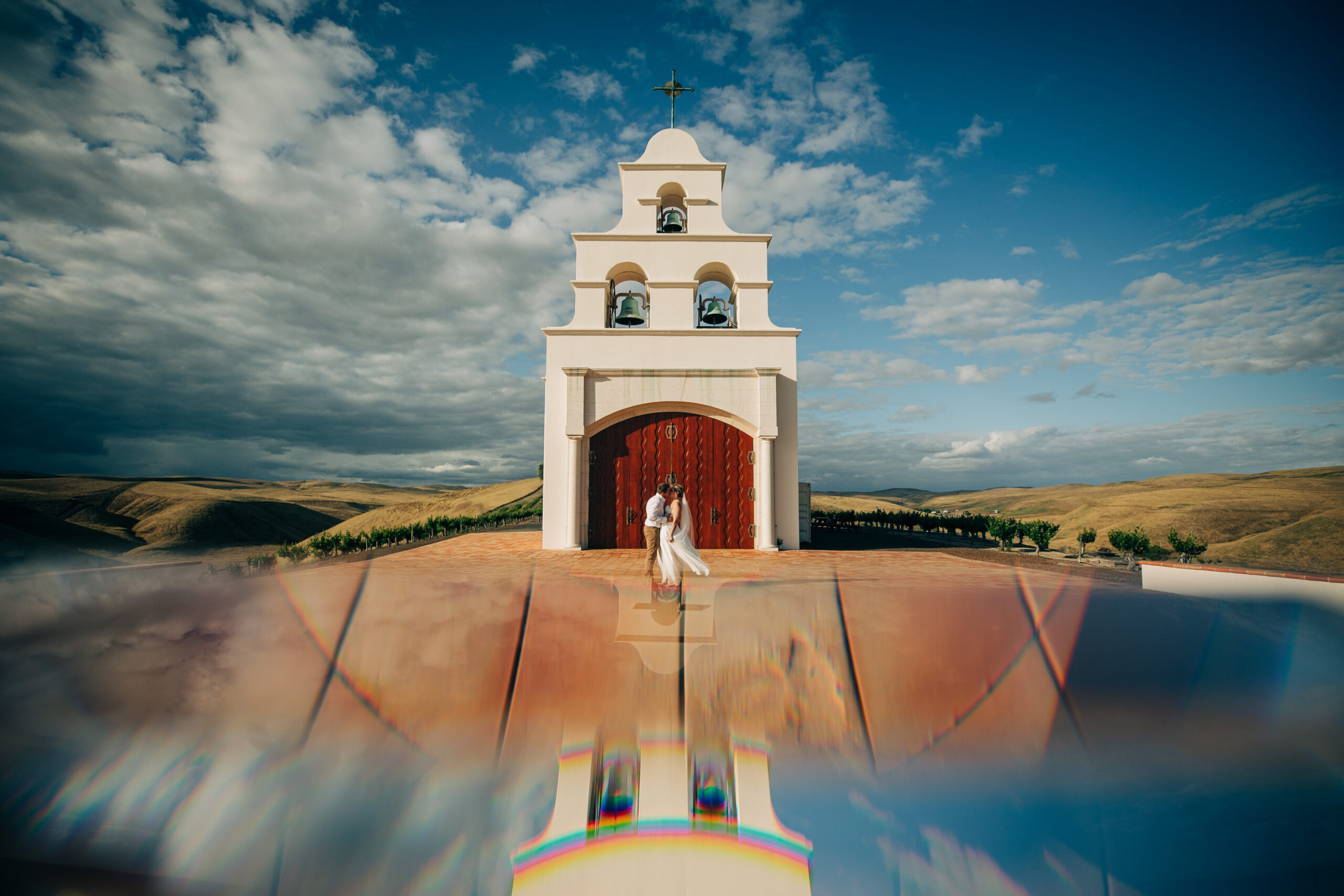 Bride and groom stand before a chapel with a dramatic reflection on the tiled courtyard.