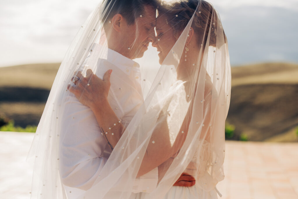 Bride and groom embrace beneath the bride's pearl-detailed wedding veil at sunset.