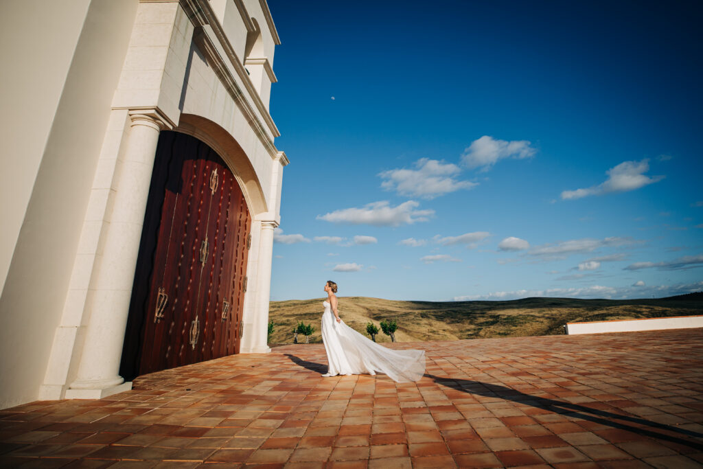 Bride stands beside the chapel with her dress flowing across the courtyard.