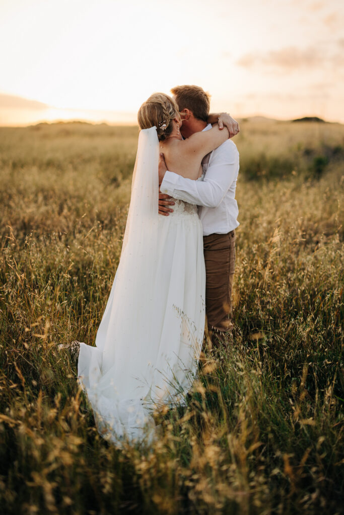 Bride and groom embracing in a golden field during sunset with the bride's veil flowing behind her.