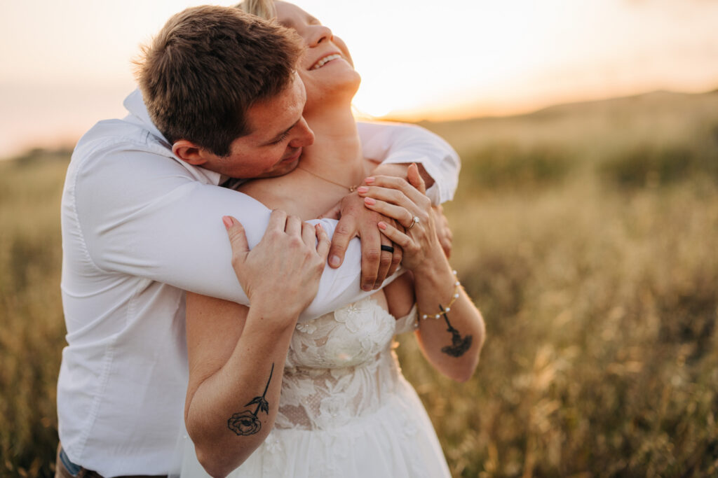 Groom embracing the bride from behind as she tilts her head back laughing in a golden meadow at sunset.