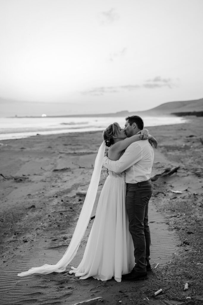 Black and white portrait of bride and groom kissing on a quiet beach at sunset.