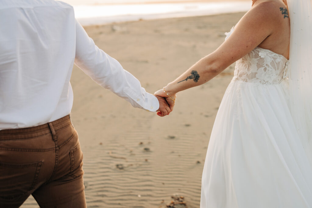 Close-up of bride and groom holding hands while walking along the beach at sunset.