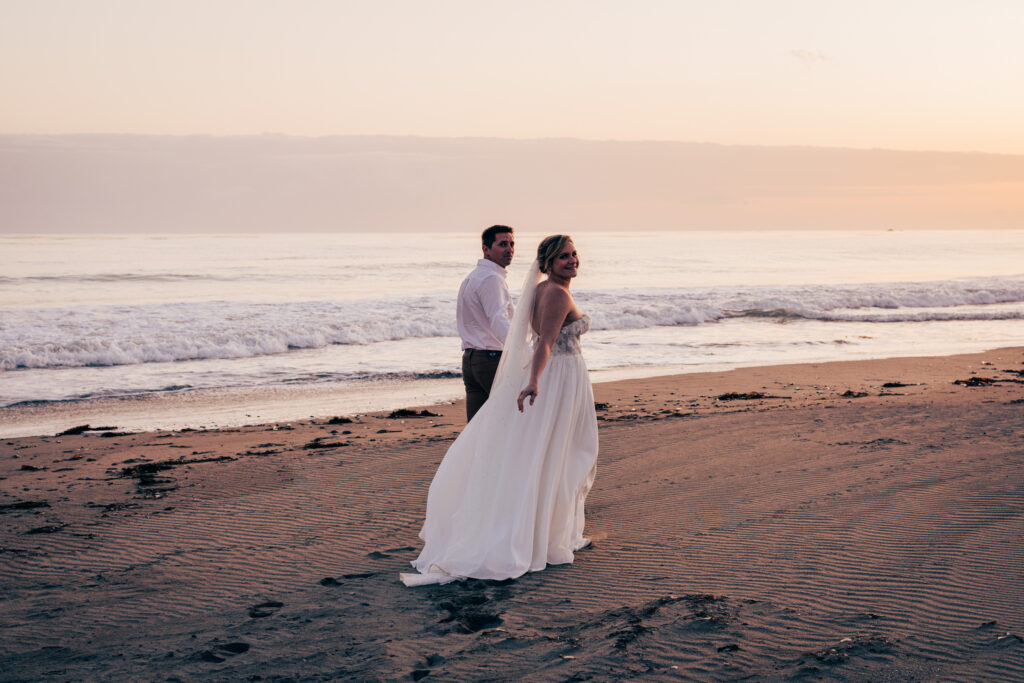 Bride and groom walking along the shoreline at sunset with waves rolling onto the beach.