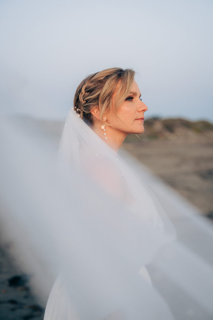 Side profile bridal portrait framed by a flowing veil on San Simeon Beach at sunset.