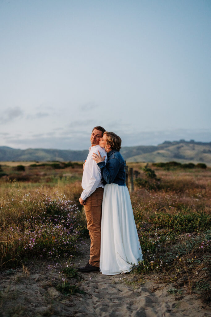 Bride kissing groom on the cheek as they stand among the dunes with rolling hills in the background.