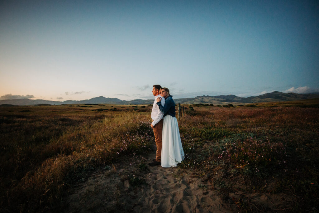 Bride and groom standing on the dunes as the sunsets, bride embracing groom from behind as she rests her cheek on his shoulder.