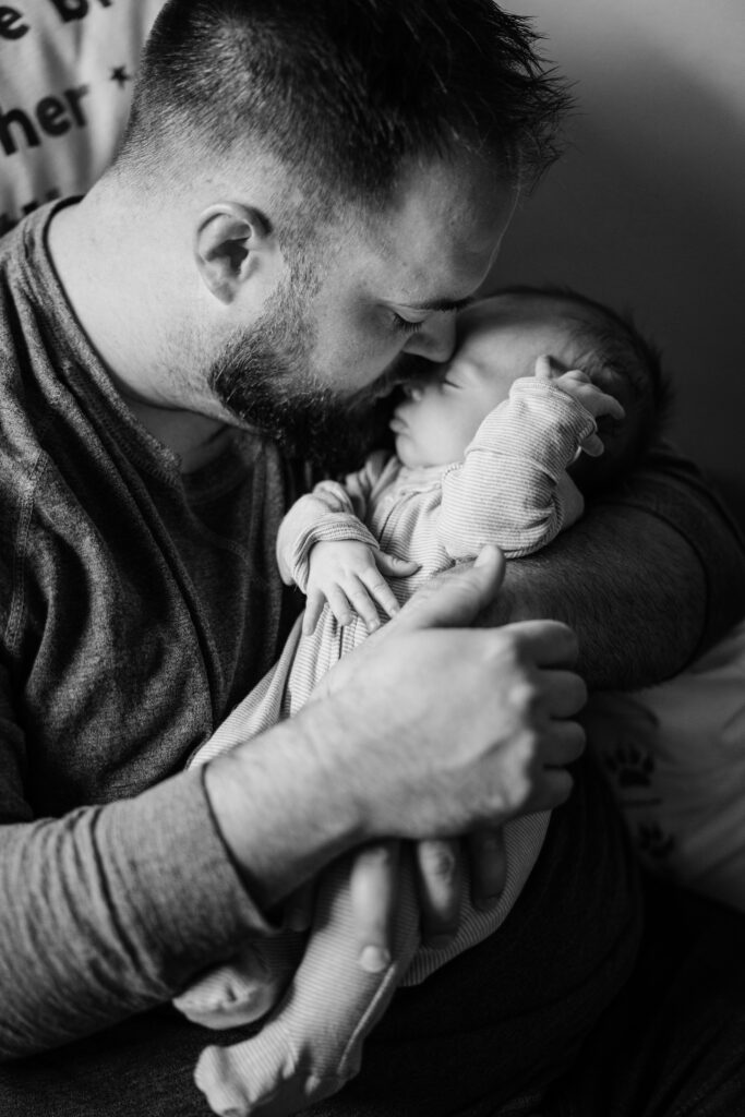 Father cradles his sleeping newborn close while resting together in soft window light.