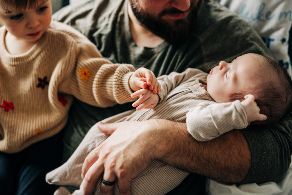 Close up of older sibling holding a sleeping newborn's hand while father watches from a nursery chair.