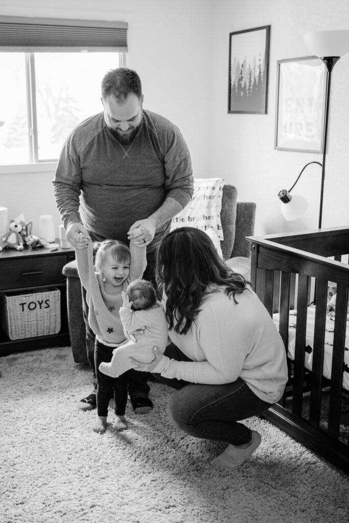 Parents play with their toddler while mother holds the newborn in a bright nursery during an in-home newborn session in Madison, Wisconsin.