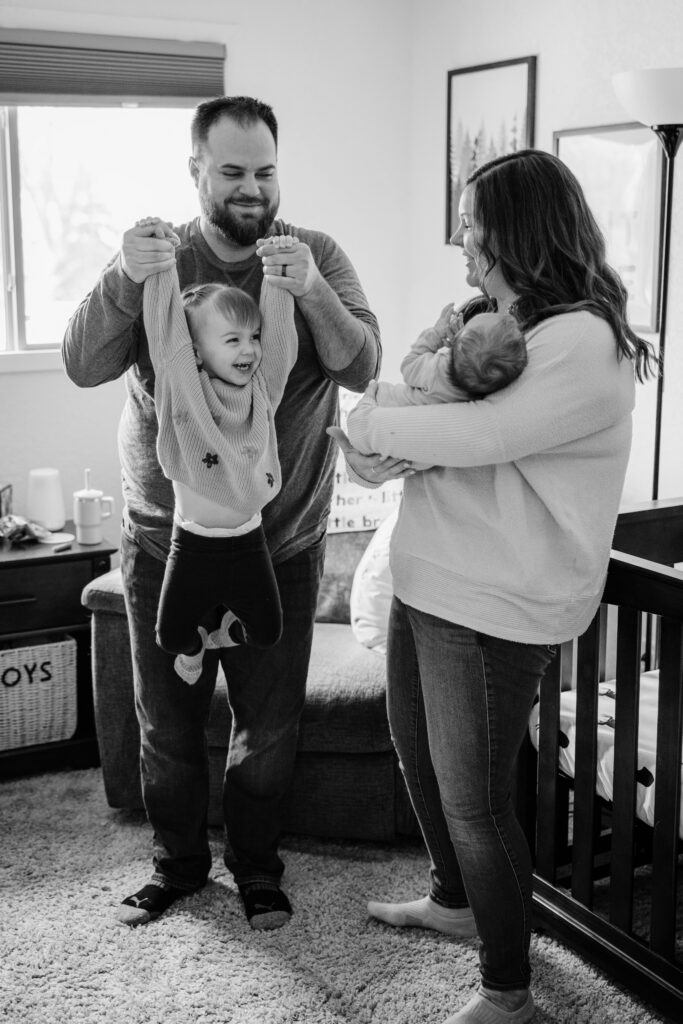 Father lifts a laughing toddler by both hands while mother cuddles the newborn beside the crib.